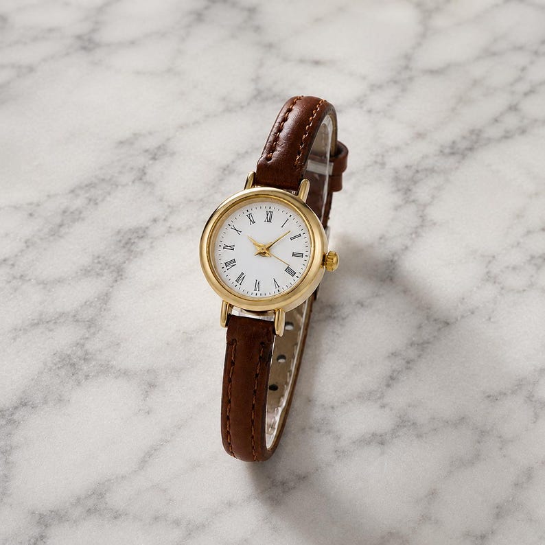 May include: A gold-tone wristwatch with a white face and Roman numeral hour markers. The watch has a brown leather band. The watch face is round and the hands are gold. The watch is a classic style.