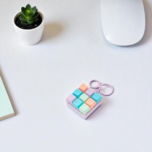 May include: A pastel-colored keycap keychain with nine square keycaps in shades of pink, orange, blue, and green. The keychain has a lavender-colored frame and a matching ring. A small succulent in a white pot and a white computer mouse are also visible.