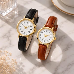 May include: Two gold-toned wristwatches with white faces and Roman numeral hour markers. One watch has a black leather band, the other a brown leather band. Both watches have gold hands and a quartz movement.