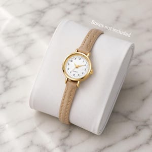May include: A gold-toned quartz watch with a white face and beige leather band. The watch has a round face with black numerals and gold hands. The watch is displayed on a white, curved surface. The text "Boxes not included" is visible.