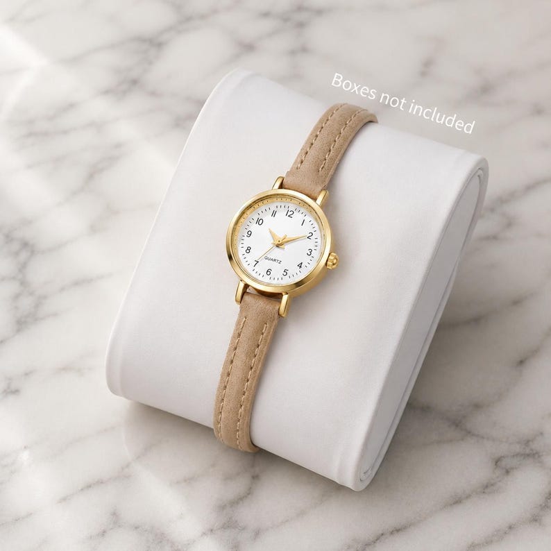May include: A gold-toned quartz watch with a white face and beige leather band. The watch has a round face with black numerals and gold hands. The watch is displayed on a white, curved surface. The text "Boxes not included" is visible.
