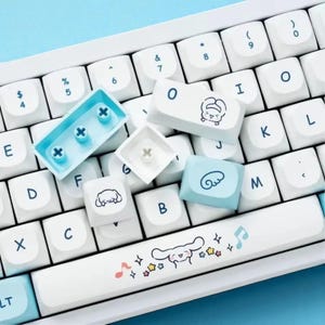 Puede incluir: Teclado blanco con teclas personalizadas con personajes de dibujos animados y notas musicales. La barra espaciadora tiene una ilustraci&oacute;n de personaje con notas musicales y estrellas. Algunas teclas son de color azul claro.