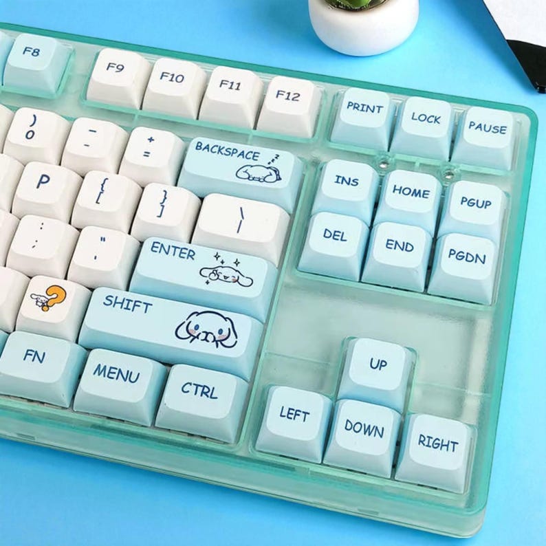 Puede incluir: Un teclado mec&aacute;nico personalizado con teclas blancas y azul claro. Las teclas tienen varios s&iacute;mbolos y texto, incluyendo "BACKSPACE", "ENTER" y teclas de flecha. Algunas teclas presentan ilustraciones de dibujos animados. El teclado est&aacute; alojado en una carcasa verde azulado transparente.