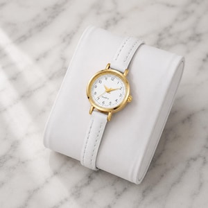May include: A gold-tone quartz watch with a white face and a white leather strap. The watch is displayed on a white, rectangular watch holder. The watch face has black numerals and gold hands.