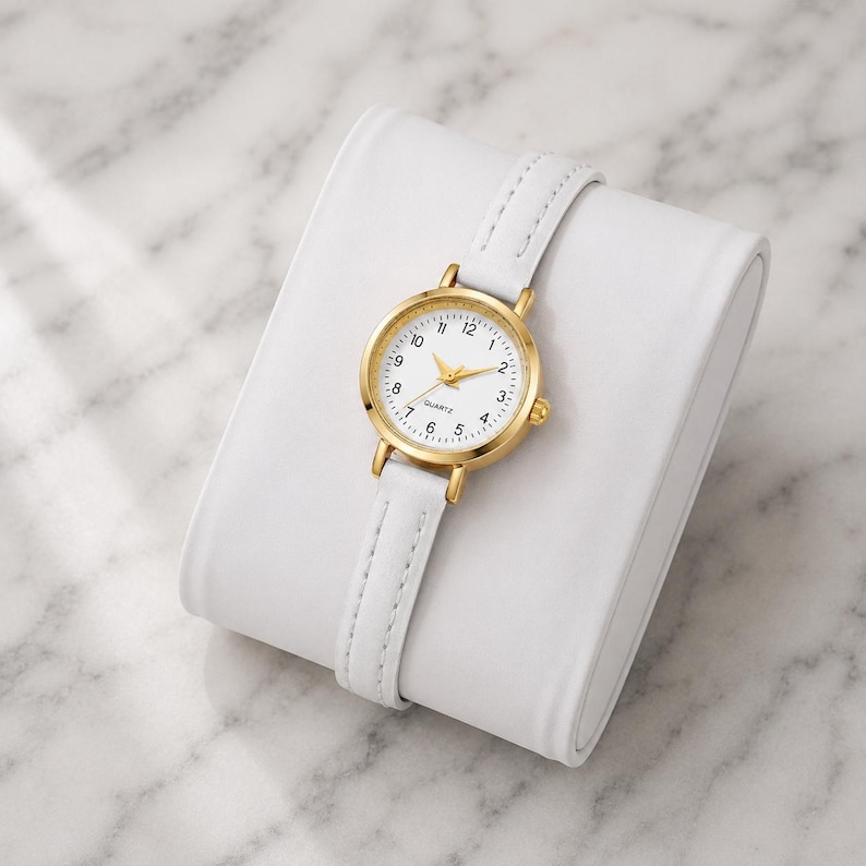 May include: A gold-tone quartz watch with a white face and a white leather strap. The watch is displayed on a white, rectangular watch holder. The watch face has black numerals and gold hands.
