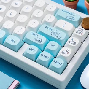 Puede incluir: Un teclado mec&aacute;nico blanco y azul claro con teclas personalizadas. Las teclas presentan ilustraciones de dibujos animados y texto como "CTRL", "SHIFT", "ENTER" y flechas direccionales. El teclado est&aacute; sobre una superficie azul.