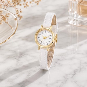 May include: A gold-toned wristwatch with a white face and Roman numeral hour markers. The watch has a white leather strap. The watch is displayed on a marble surface.