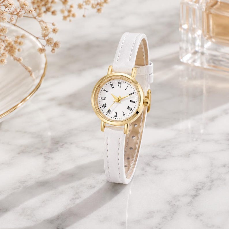 May include: A gold-toned wristwatch with a white face and Roman numeral hour markers. The watch has a white leather strap. The watch is displayed on a marble surface.
