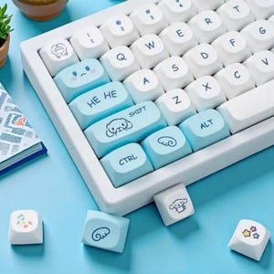 Puede incluir: Un teclado mec&aacute;nico blanco y azul claro con teclas personalizadas. Las teclas presentan ilustraciones de dibujos animados y frases como "HE HE" y "SHIFT". El teclado est&aacute; sobre una superficie azul claro.