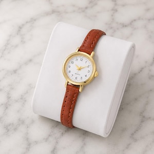 May include: A gold-tone quartz watch with a white face and brown leather strap. The watch has a round face with black numerals and gold hands. The strap is a rich brown color and appears to be made of leather. The watch is displayed on a white surface.