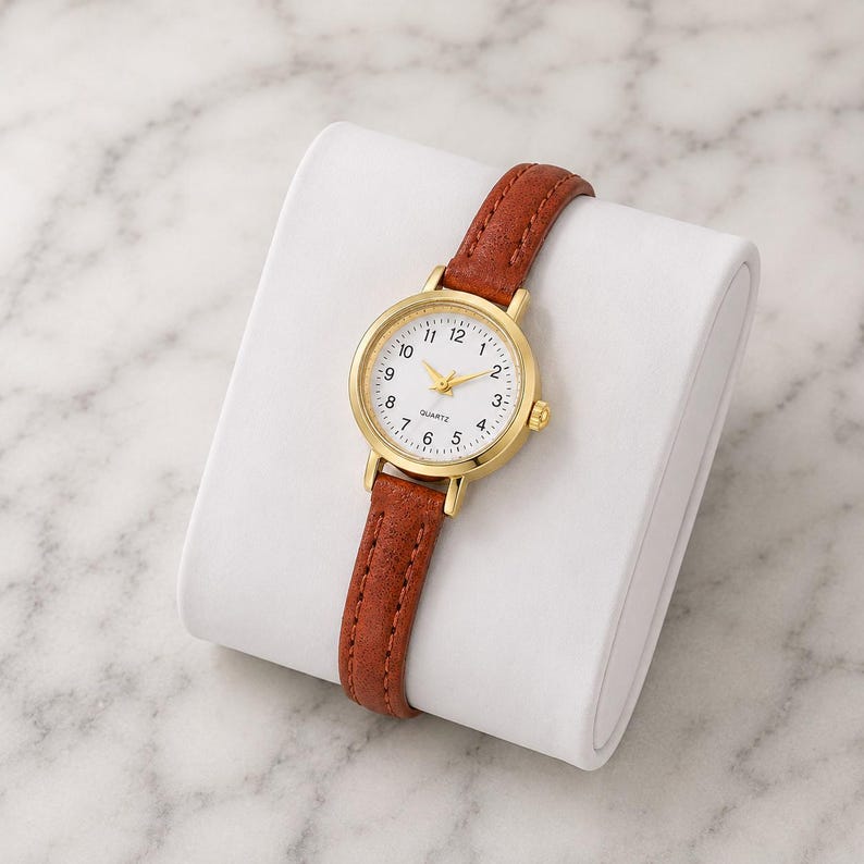 May include: A gold-tone quartz watch with a white face and brown leather strap. The watch has a round face with black numerals and gold hands. The strap is a rich brown color and appears to be made of leather. The watch is displayed on a white surface.
