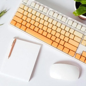 May include: A white mechanical keyboard with gradient orange keycaps, a white mouse, a rose gold pen, and a blank white notepad. A small potted plant and sprig of rosemary are also visible.