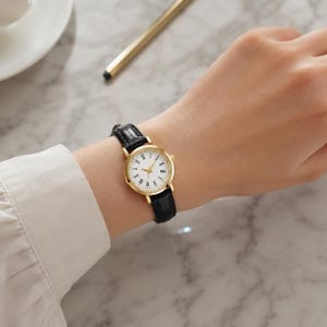 May include: A gold-toned wristwatch with a white face and black Roman numerals. The watch has a black leather band. The watch is on a wrist.