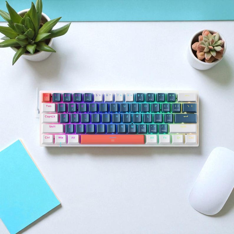 May include: A white mechanical keyboard with a rainbow gradient keycap design. The keyboard has a rectangular shape with a variety of keys, including function keys, letters, and numbers. The keyboard is surrounded by a white mouse, a blue notepad, and small potted plants.
