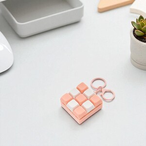 May include: A square keychain with a grid pattern of pink and white squares. The keychain has a pink border and a matching pink keyring. The background is a white surface with a few other objects.
