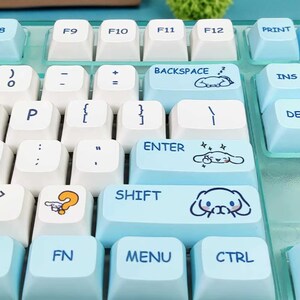 Puede incluir: Primer plano de un teclado mec&aacute;nico con teclas en azul claro y blanco. Las teclas tienen varios s&iacute;mbolos y texto, incluyendo "BACKSPACE", "ENTER" y "SHIFT". Algunas teclas presentan ilustraciones de personajes de dibujos animados.
