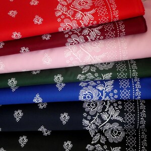 Op de afbeelding: Een stapel kleurrijke stofstalen in rood, bordeaux, roze, groen, blauw en zwart. Elke stof heeft een witte bloemen- en geometrische rand. De stoffen zijn gevouwen en gelaagd, wat de verscheidenheid aan kleuren en patronen laat zien.