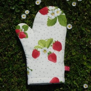 Puede incluir: Manopla de horno blanca con estampado de fresas. La manopla presenta fresas rojas, hojas verdes y flores blancas sobre un fondo de pequeños lunares grises. Se adjunta una pequeña cinta de lunares rojos y blancos. La palabra "Delicious" está impresa.
