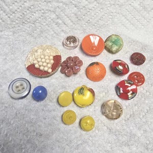 May include: A collection of colorful vintage buttons in various shapes and sizes. Includes a blue button, a round orange button, a flower-shaped button, and a button with a grape design. The buttons are arranged on a white surface.
