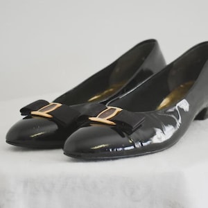 May include: Pair of black patent leather shoes with gold and black bows. The shoes have a low heel.