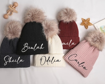 May include: Five personalized knit beanies with faux fur pom-poms. Colors include charcoal, black, cream, burgundy, and pink. Each beanie has a name embroidered in a cursive font. A wooden rattle and star-shaped decoration are also visible.