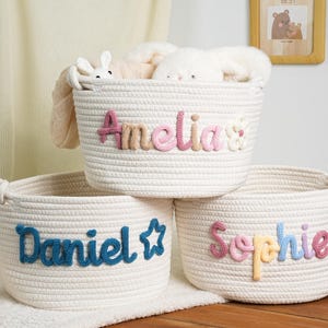 Könnte beinhalten: Drei weiße Seilkörbe mit personalisierten Namen: "Amelia", "Daniel" und "Sophie". Die Namen sind in bunten, gestickten Buchstaben. Die Körbe sind mit weichen Spielzeugen gefüllt, ideal für ein Kinderzimmer.