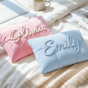 Puede incluir: Dos cojines rectangulares, uno rosa y otro azul claro, con los nombres "Olivia" y "Emily" en escritura cursiva. Los cojines están sobre una colcha blanca, con un portátil y una revista en el fondo.