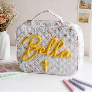 Puede incluir: Un bolso acolchado con estampado floral con un diseño "Bella" de ganchillo amarillo y una cruz. El bolso tiene un asa y está sobre una superficie blanca. También hay un jarrón con flores, bolígrafos y palitos de colores.