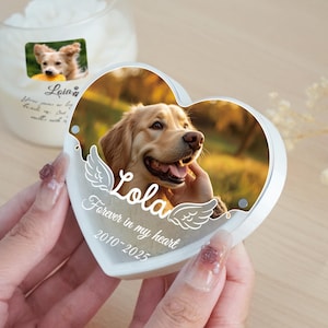 May include: A heart-shaped memorial featuring a photo of a golden retriever, the name "Lola", and the text "Forever in my heart 2010-2025". The memorial is white with a clear border and angel wing accents. A candle with a dog photo is in the background.