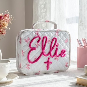 May include: White quilted cosmetic bag with pink butterfly accents and the name "Ellie" in a bold, pink, embroidered script. The bag has a handle and a cross symbol. A vase of dried flowers and a teacup are in the background.