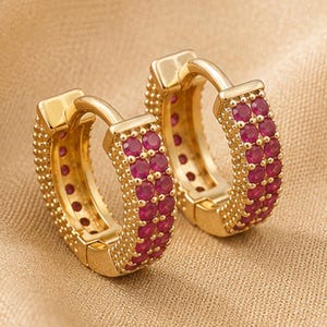 May include: A pair of gold hoop earrings featuring two parallel rows of small, round, vibrant red gemstones set along the front face, with a textured, beaded gold finish on the outer edges of the hoops and a secure hinged closure mechanism.