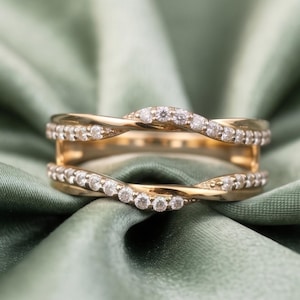 May include: A gold ring guard featuring two curved, interlocking bands that meet in the center. Each band is set with a row of small, clear, round-cut stones along the outer edge, creating a continuous sparkling effect across the metal surface.