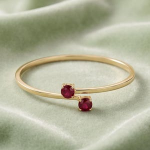 Puede incluir: Un anillo abierto de oro con dos rubíes de color rojo intenso en talla redonda, engastados en los extremos de la banda fina y pulida. El diseño presenta una silueta abierta donde las dos piedras preciosas vibrantes se superponen ligeramente.