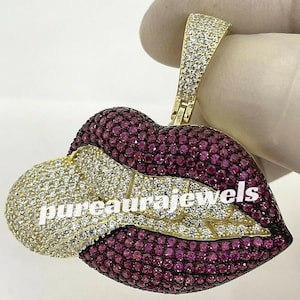 May include: A gold-toned pendant in the shape of lips, encrusted with small, round, ruby-colored and clear gemstones. The pendant has the text "pureaurajewels" in white. The bail is also encrusted with clear gemstones.