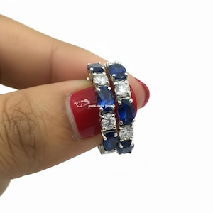 May include: Two silver-toned eternity hoop earrings. Each earring features alternating oval and round blue gemstones and clear, round gemstones. The earrings are held by a person with red nail polish.