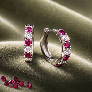 May include: A pair of silver-toned hoop earrings featuring an alternating pattern of round-cut vibrant red rubies and clear white diamonds. Several loose, round-cut red rubies are scattered nearby, showcasing their deep crimson colour and faceted surface.