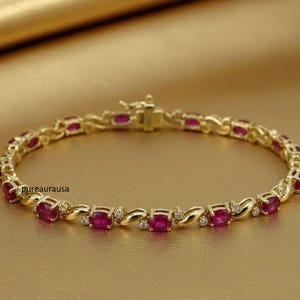 Puede incluir: Pulsera dorada con piedras preciosas de rubí de corte ovalado y pequeños detalles de diamantes. La pulsera presenta un diseño delicado e entrelazado y un cierre seguro. Las piedras preciosas son de color rojo vibrante.