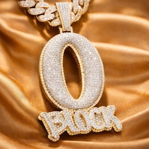 Puede incluir: Un collar con colgante dorado con la letra "O" y la palabra "BLOCK" incrustadas con piedras preciosas brillantes. El collar tiene una cadena gruesa y descansa sobre una superficie de satén dorado.