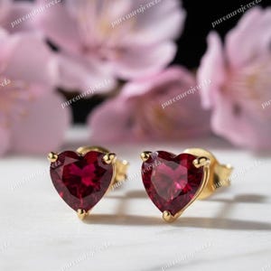 Puede incluir: Un par de pendientes de botón en forma de corazón. Cada pendiente presenta una gema roja vibrante engastada en un metal dorado. Las gemas están talladas para parecer un corazón. Los pendientes se muestran sobre un fondo floral suave y borroso.