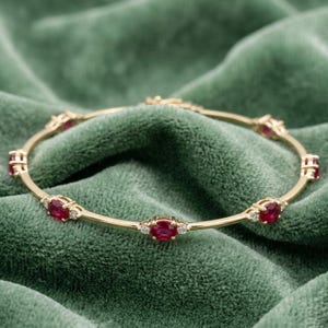 May include: A gold bangle bracelet featuring a series of oval-cut red rubies spaced evenly along the band. Each ruby is flanked by two small, clear, round-cut accent stones set into the gold metal, creating a repeating pattern around the circular frame.