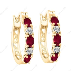 Könnte beinhalten: Ein Paar goldene Creolen. Jeder Ohrring hat drei ovale, tiefrote Edelsteine und zwei runde, klare Edelsteine. Die Ohrringe haben ein klassisches Design mit einem sicheren Verschluss.