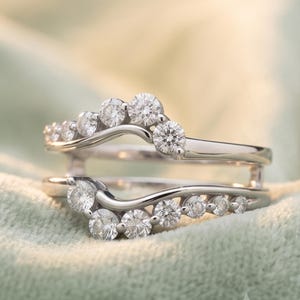 May include: A silver-toned metal ring guard featuring two curved bands connected at the sides. The bands are adorned with a series of small, round, clear faceted stones set along the top edges, creating a shimmering, arched, and layered appearance.