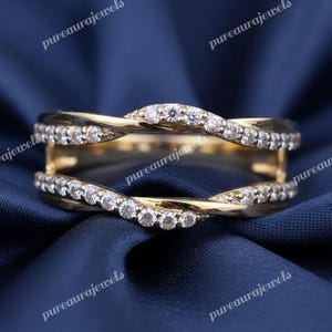 May include: A gold ring featuring a double band design, with the bands interwoven and set with small, clear gemstones. The ring is displayed against a dark blue fabric background, which accentuates the ring's intricate details.