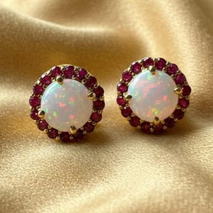 May include: A pair of round stud earrings featuring a central cabochon opal with iridescent flashes of colour, surrounded by a halo of small, deep red rubies set in yellow gold. The circular design highlights the contrast between the white opal and red stones.
