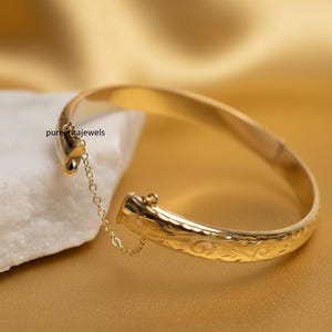 May include: A gold bangle bracelet with an engraved floral pattern. The bracelet is open, showing a small chain linking the two sides. The background is a golden-yellow colour.