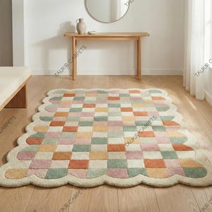 May include: A multi-coloured, checkered rug with a scalloped edge. The rug features squares of pastel colours including pink, orange, green, and cream. The rug is placed on a wooden floor in a room with a bench and a console table.