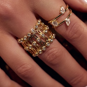 Può includere: Due anelli d'oro con pietre preziose trasparenti. Un anello è una fascia larga con un design a griglia e piccole pietre rotonde. L'altro anello ha una doppia fascia con una pietra a forma di pera e una pietra rotonda.