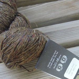 May include: A skein of brown yarn with flecks of red, yellow, and blue. The yarn is labeled "Wood 835" and "Lot: 36115". The label also includes instructions for washing and drying the yarn, as well as information about the yarn's origin: "Made in Argentina".
