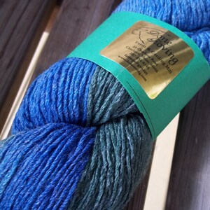 K&ouml;nnte beinhalten: Ein Kn&auml;uel blauer und gr&uuml;ner Wolle mit einem goldenen Etikett, auf dem "Done Zoving Farm & Carding Mill" mit Kontaktinformationen steht.