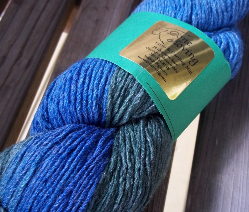 K&ouml;nnte beinhalten: Ein Kn&auml;uel blauer und gr&uuml;ner Wolle mit einem goldenen Etikett, auf dem "Done Zoving Farm & Carding Mill" mit Kontaktinformationen steht.